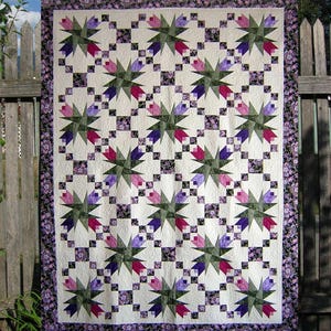 Op de afbeelding: Een handgemaakte quilt met een bloemenpatroon. De quilt heeft een herhalend ontwerp van gestileerde bloemen in tinten paars, roze en groen tegen een witte achtergrond. De rand is een bloemenprint in paars en wit. De quilt wordt buiten tentoongesteld.