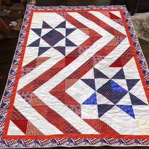 May include: A patriotic quilt featuring a red, white, and blue color scheme. The quilt showcases a star pattern with red and white striped and dotted fabric, and blue star accents. The border is a repeating pattern of stars and stripes.
