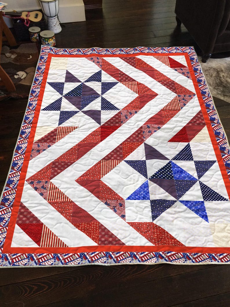 May include: A patriotic quilt featuring a red, white, and blue color scheme. The quilt showcases a star pattern with red and white striped and dotted fabric, and blue star accents. The border is a repeating pattern of stars and stripes.