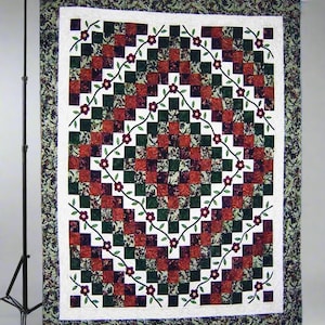 Peut inclure: Une courtepointe faite à la main avec un motif central en losange de carrés rouges et verts. Des vignes florales avec des fleurs violettes décorent la bordure blanche. La courtepointe est encadrée par une bordure à motifs dans des tons de vert et de bordeaux.