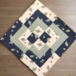 Könnte beinhalten: Ein quadratischer Quilt mit einem Rautenmuster. Der Quilt hat abwechselnde Bänder aus cremefarbenen, marineblauen und hellgrünen Stoffen. Die cremefarbenen und marineblauen Stoffe sind mit einem Muster aus Eicheln und Blättern verziert. Der Quilt ist aus Stoff.
