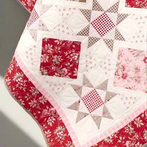 Peut inclure: Une couverture matelassée à motifs floraux rouges, roses et blancs. La couverture présente des motifs d'étoiles et de carrés, avec une bordure florale rouge. La couverture est en tissu doux et présente un motif de couture détaillé.