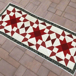May include: A rectangular quilt featuring a star pattern in shades of red and white. The stars are set against a cream background, with a black and green floral border. The quilt is likely made of cotton.