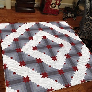 Puede incluir: Un edredón hecho a mano con un patrón geométrico. El edredón presenta un fondo blanco con bloques grises y grises oscuros dispuestos en un patrón diagonal. Estrellas rojas están espaciadas uniformemente en el diseño. El edredón es probablemente de algodón.