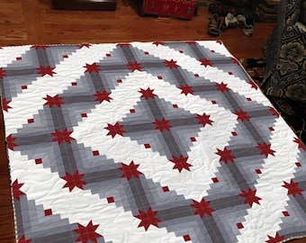 May include: A handmade quilt with a geometric pattern. The quilt features a white background with gray and dark gray blocks arranged in a diagonal pattern. Red star shapes are evenly spaced throughout the design. The quilt is likely made of cotton.