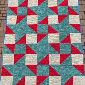 以下が含まれることがあります： 幾何学模様の手作りキルト。赤、白、ティール色の正方形と三角形が交互に配置されています。青い縁取りがあり、おそらく綿製です。約150cm x 180cmです。