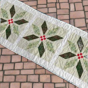 Könnte beinhalten: Ein gesteppter Tischläufer mit einem sich wiederholenden Sternmuster. Das Design enthält grüne, braune und rote Akzente auf cremefarbenem Hintergrund. Der Läufer hat einen weißen Rand mit einem Steppmuster.