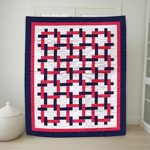 May include: A rectangular quilt with a woven pattern of red and navy blue rectangles on a white background. The quilt has a navy blue border and a red dotted inner border. The quilt is hanging on a white door.