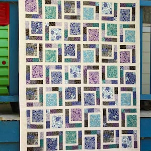 May include: A handmade quilt with a repeating pattern of squares in shades of blue, purple, and teal, framed by dark brown and white borders. The quilt is displayed outdoors, showcasing its intricate patchwork design.