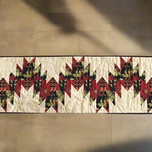 May include: A rectangular quilted table runner with a cream background and a repeating pattern of red poinsettias and green leaves on a black background. The runner is bordered with a black trim.