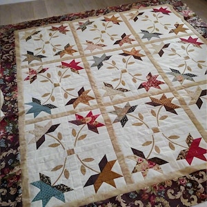 May include: A handmade quilt featuring a repeating star pattern in various colors, including red, brown, and blue, set against a cream background. The quilt is bordered with a floral patterned fabric. The quilt is approximately 70 inches by 90 inches.