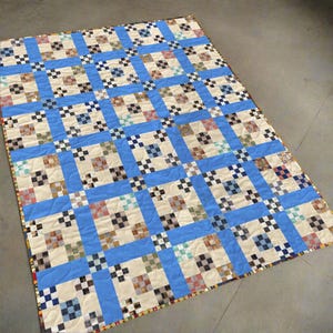 Op de afbeelding: Een handgemaakte quilt met een rasterpatroon, met vierkanten in verschillende kleuren en kleine zwart-wit geblokte vierkanten. Het heeft lichtblauwe randen en een crèmekleurige achtergrond. De randen zijn afgewerkt met een patroonrand.