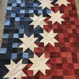 May include: A handmade quilt with a patriotic theme. The quilt features a design of alternating red and blue squares, with large white star shapes. The colours are arranged to create a flag-like pattern, ideal for home decor.