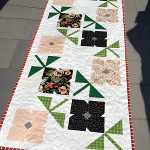 Peut inclure: Chemin de table matelassé blanc rectangulaire avec un motif floral. Le chemin de table présente des blocs floraux alternés en noir, pêche et tissus à motifs, accentués de triangles verts. Les bords sont bordés d'une bordure rayée rouge et blanche.