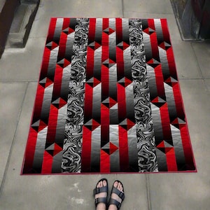 May include: A rectangular quilt with a geometric pattern in red, black, gray, and white. The design features vertical stripes and triangular accents. The quilt is bordered in red.