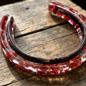 May include: A horseshoe-shaped cuff bracelet made of clear resin. The resin is embedded with vibrant red fragments and metallic gold flakes. A smooth, dark, curved band runs along the inner arc of the clear resin structure, creating a layered effect.