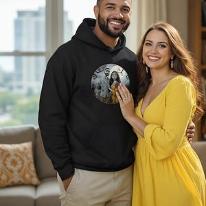 Peut inclure: Un sweat &agrave; capuche noir avec un motif circulaire repr&eacute;sentant un couple de dessins anim&eacute;s. Une femme porte une robe jaune. L'image est prise &agrave; l'int&eacute;rieur avec une fen&ecirc;tre et un canap&eacute; en arri&egrave;re-plan.