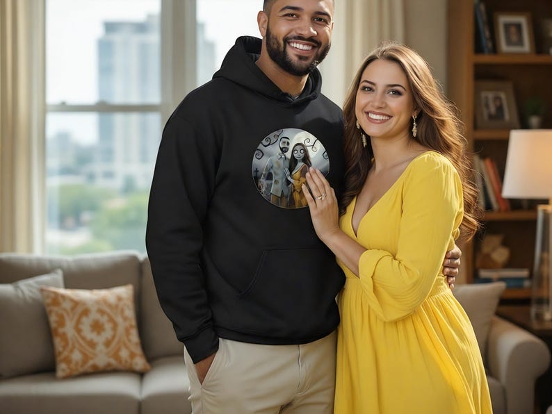 Peut inclure: Un sweat &agrave; capuche noir avec un motif circulaire repr&eacute;sentant un couple de dessins anim&eacute;s. Une femme porte une robe jaune. L'image est prise &agrave; l'int&eacute;rieur avec une fen&ecirc;tre et un canap&eacute; en arri&egrave;re-plan.