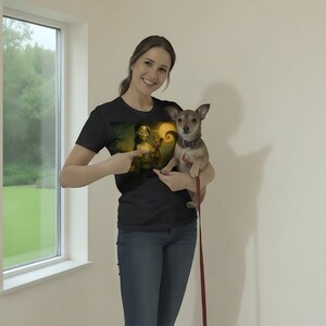 May include: Black t-shirt with a graphic design featuring a character with a yellow light and a swirling background. The person is holding a small brown dog with a leash. The person is pointing at the graphic on the shirt.