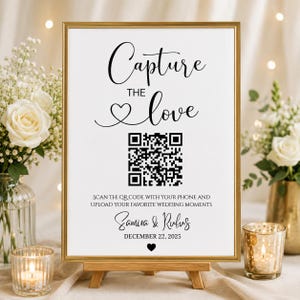 Könnte beinhalten: Ein weißes rechteckiges Schild in einem goldenen Rahmen auf einer Holzstaffelei. Der Text lautet Capture the love mit einem Herzsymbol, einem QR-Code und der Anweisung Scan the QR code with your phone and upload your favorite wedding moments. Darunter stehen die Namen Samira & Rufus, das Datum 22. Dezember 2025 und ein kleines Herz.