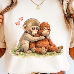 May include: A cream-coloured t-shirt with a charming illustration of two monkeys embracing. One is light brown, the other reddish-brown. Pink hearts float above them, and small flowers are scattered on the green grass. A sweet design.