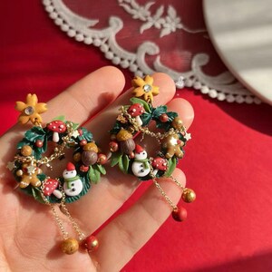 May include: A pair of festive earrings featuring miniature Christmas wreath designs. Each wreath is adorned with green leaves, red mushrooms, snowmen, gingerbread men, and gold accents. The earrings dangle with gold chains and red and gold beads.