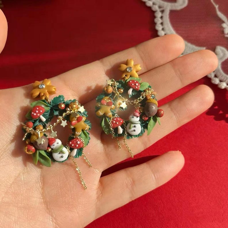 May include: A pair of festive earrings featuring miniature wreaths with green leaves, red and white mushrooms, gingerbread men, snowmen, and gold accents. Each wreath is adorned with a yellow flower and hangs from a delicate gold chain.