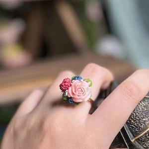 以下が含まれることがあります： ミニチュアの花束が特徴のゴールドトーンのリング。リングの焦点は、繊細なピンクのバラで、赤いベリー、青いアクセント、緑の葉で補完されています。リングは指に着用されています。