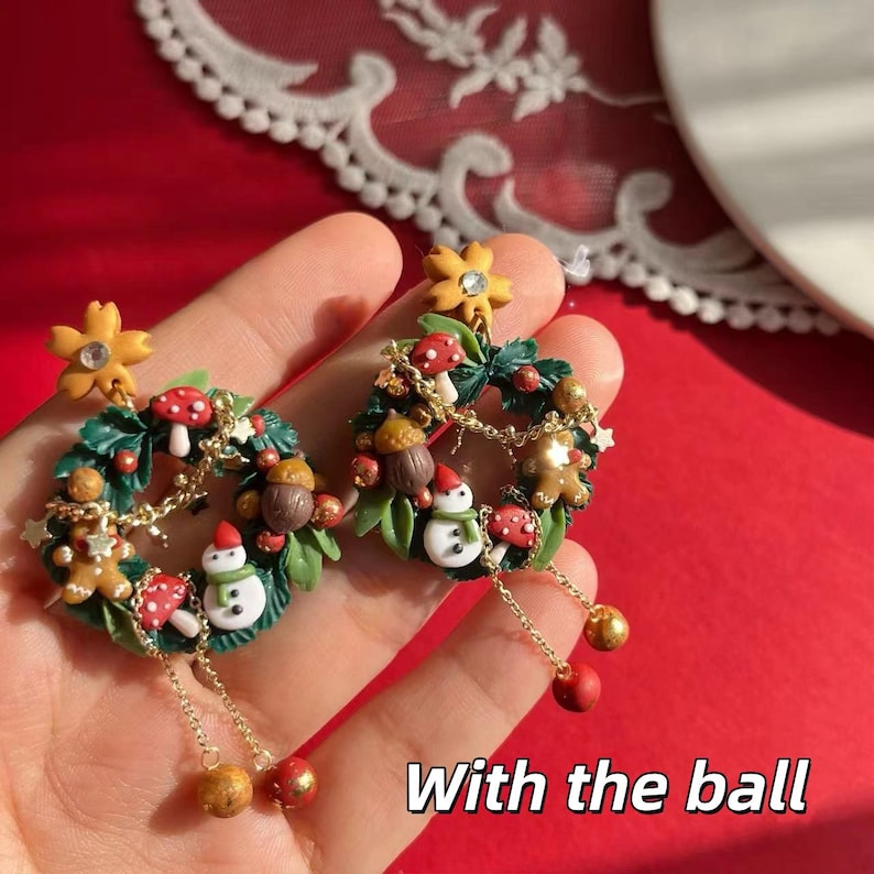 May include: A pair of festive earrings featuring miniature Christmas wreath designs. Each earring has a green wreath adorned with red mushrooms, gingerbread men, snowmen, and gold accents. The earrings dangle with gold chains and red and gold ball charms. The text "With the ball" is visible.