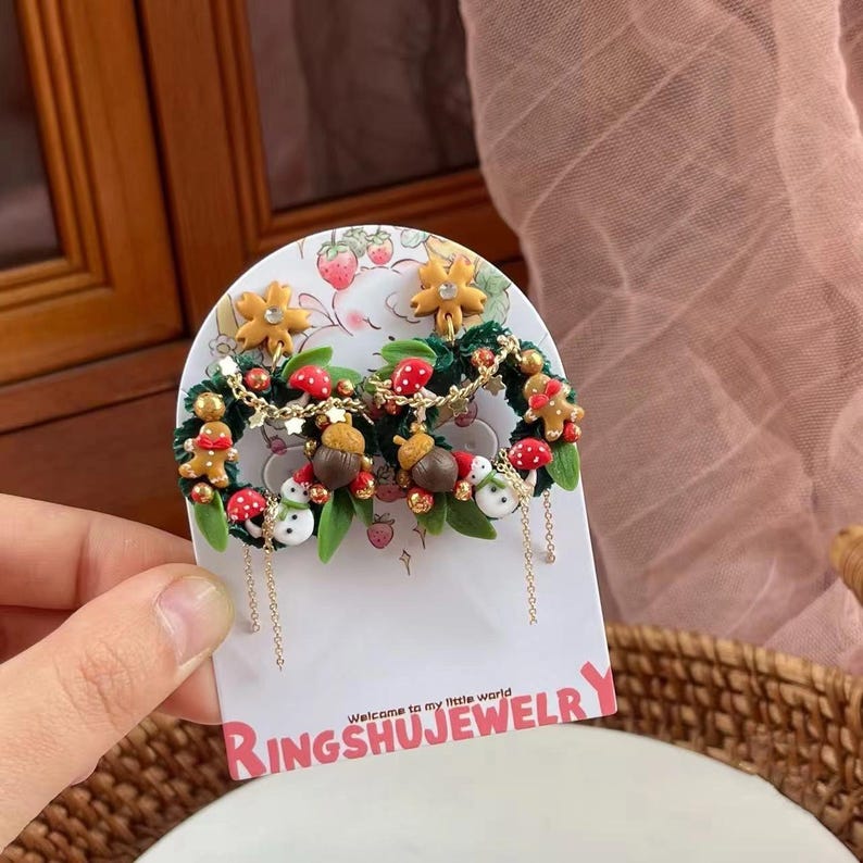 May include: A pair of festive earrings featuring miniature wreaths with gingerbread men, mushrooms, acorns, and snowmen. The earrings are adorned with gold-colored chains and are displayed on a card with the text "RingshuJewelry".