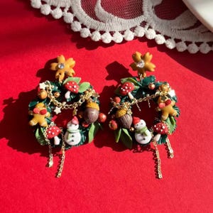 May include: A pair of festive Christmas wreath earrings. Each earring features a miniature wreath with a snowman, gingerbread man, mushrooms, and acorns. The earrings are adorned with gold-colored chains and a yellow flower at the top, set against a red background.