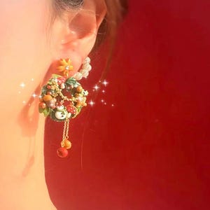 May include: A festive earring featuring a miniature wreath design with a snowman, mushrooms, and acorns. The wreath is adorned with green leaves, red berries, and a gold chain with dangling ornaments. The earring has a yellow flower and white accents.