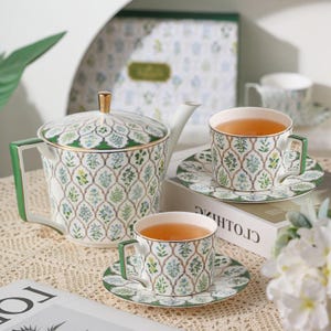 Peut inclure: Service à thé en céramique blanche avec un motif floral vert, bleu et doré. L'ensemble comprend une théière, deux tasses et des soucoupes. Les tasses sont remplies de thé. La théière a une poignée et un couvercle dorés.