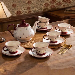 Puede incluir: Un juego de té de cerámica blanca con una tetera, seis tazas y platillos. El juego presenta un estampado floral con detalles en rojo y azul. Cucharas doradas dispuestas a un lado. La tetera tiene tapa y asa burdeos.