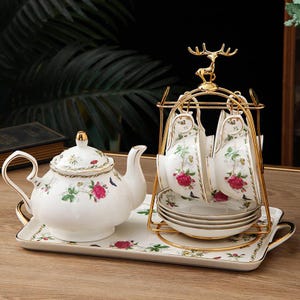 Puede incluir: Juego de té de porcelana blanca con diseño floral de rosas rojas y hojas verdes. El juego incluye una tetera, tazas, platillos y una bandeja. Las tazas están suspendidas de un estante de metal dorado.
