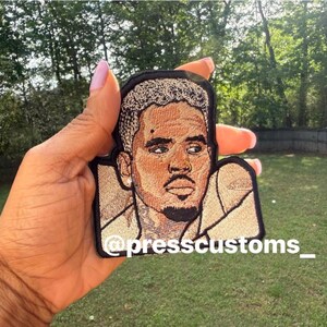 May include: Embroidered patch featuring a portrait of a man with light brown skin, light brown hair, and a goatee. The patch is held in a hand against a backdrop of green grass and trees. The text "@presscustoms_" is at the bottom.