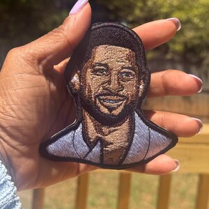 May include: Embroidered patch featuring a smiling man with dark hair and a beard. The patch has a black border and a light blue suit. The image is detailed with brown and tan thread, creating a realistic portrait.