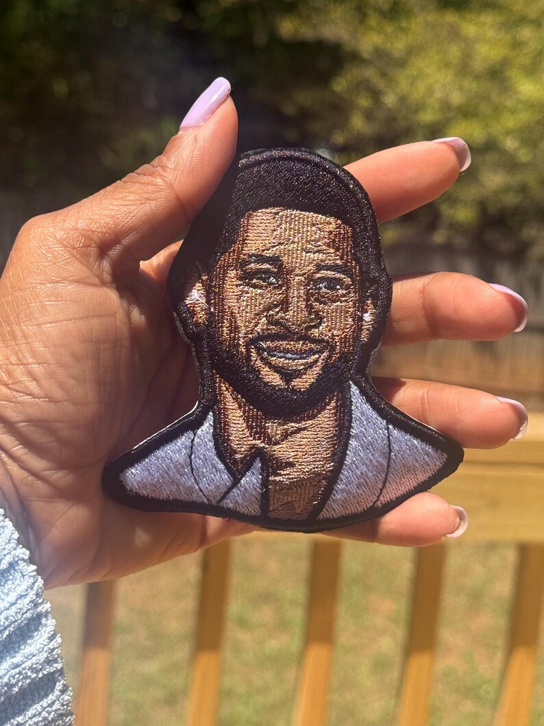 May include: Embroidered patch featuring a smiling man with dark hair and a beard. The patch has a black border and a light blue suit. The image is detailed with brown and tan thread, creating a realistic portrait.