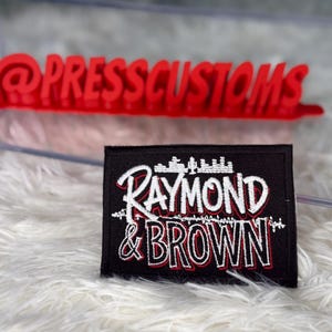 May include: Black rectangular patch with the text "RAYMOND & BROWN" in white and red lettering. The patch has a cityscape design above the text. A red "@PRESSCUSTOMS" sign is in the background.