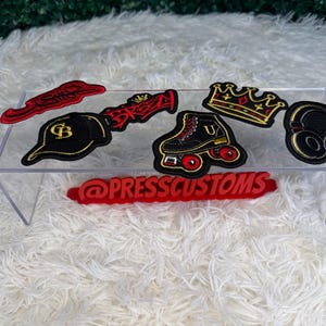 May include: A collection of embroidered patches featuring a red script logo, a black baseball cap with gold letters, a red graffiti-style logo, a black roller skate with red wheels, a gold crown, and black headphones with the text @PRESSCUSTOMS.