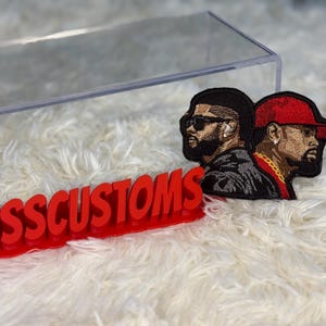 May include: Embroidered patch featuring two men in profile, one wearing sunglasses and the other a red cap. The patch is on a white, fluffy surface. The red text "PRESSCUSTOMS" is in the foreground.