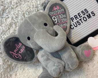 May include: A gray plush elephant with pink heart accents on its feet. The elephant's ears display custom text, including a name and birth details: date, weight (10 ounces), and length (21 inches). The sign in the background reads "PRESS CUSTOMS."