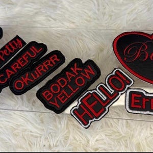 May include: A collection of black and red embroidered patches with various phrases, including "PRESS", "Pretty & Petty", "BE CAREFUL", "OKURRR", "BODAK YELLOW", "HELLO!", "Err Time", and a heart-shaped patch with "Bardi".