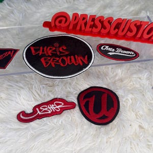 May include: A collection of custom patches in various shapes and sizes. The patches feature red and white text on black backgrounds, including the names "Chris Brown" and "Usher", and the word "breezy". A red "@PRESSCUSTOMS" sign is also present.