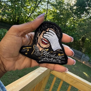 May include: Embroidered patch featuring a portrait of Michael Jackson, with his hand covering his face. The patch has a black background with gold accents and the name "Michael Jackson" in cursive. The patch is held up against a background of green trees.