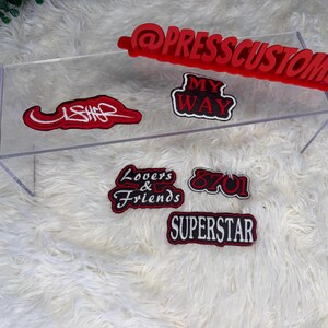 May include: A collection of red and black embroidered patches with various text designs, including "USHER," "MY WAY," "Lovers & Friends," "8701," and "SUPERSTAR." A red sign with the text "@PRESSCUSTOMS" is also present. The patches are displayed on a clear acrylic stand.