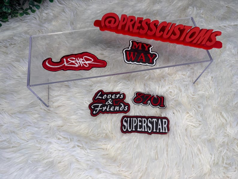 May include: A collection of red and black embroidered patches with various text designs, including "USHER," "MY WAY," "Lovers & Friends," "8701," and "SUPERSTAR." A red sign with the text "@PRESSCUSTOMS" is also present. The patches are displayed on a clear acrylic stand.