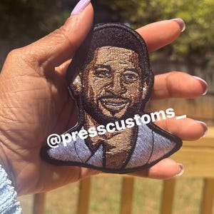 May include: Embroidered patch featuring a smiling man with a light blue suit. The patch is black around the edges and has the text "@presscustoms_". The patch is held up by a hand.