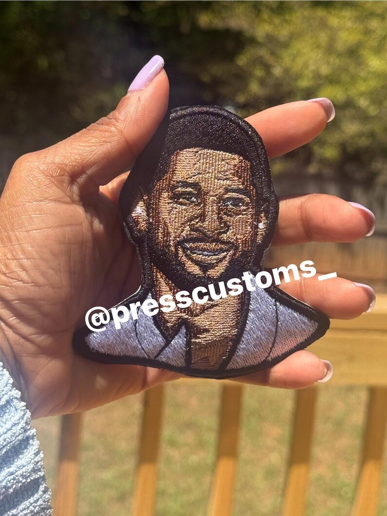 May include: Embroidered patch featuring a smiling man with a light blue suit. The patch is black around the edges and has the text "@presscustoms_". The patch is held up by a hand.