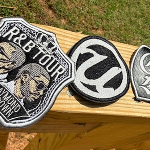 May include: Three embroidered patches. The largest is shield-shaped with "R&B TOUR" and "Usher Raymond & Chris Brown" text. A black and white oval patch and a heart-shaped patch with stylized letters complete the set.